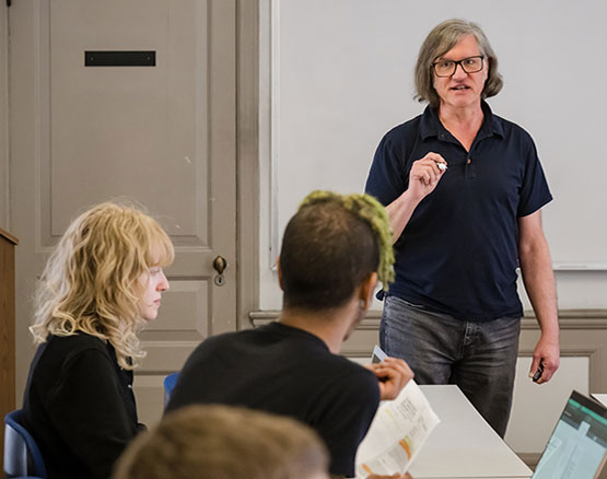 A professor teaching a class at the front of a classroom with students.