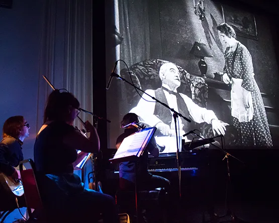 A small live ensemble—featuring a guitarist, a violinist, and a pianist—performing in front of a large projection screen showing a black-and-white film scene of a man seated in an armchair and a woman standing in a polka-dot dress.