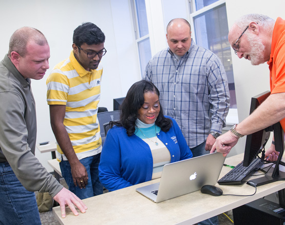 Several students work on computer with professor pointing out something on screen