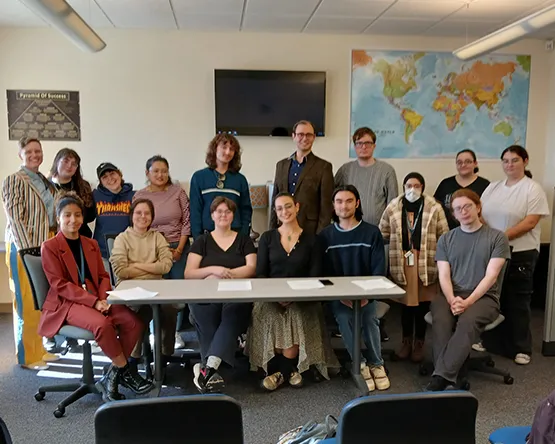 Two rows of students and faculty pose together at table with world map on wall behind them.