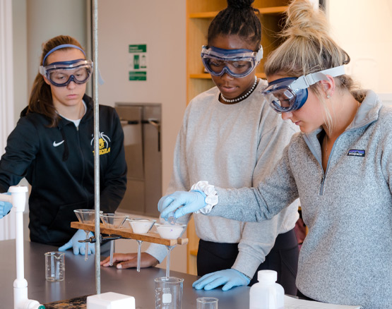 Three Webster students wearing gloves and goggles work with chemicals in funnels and beakers