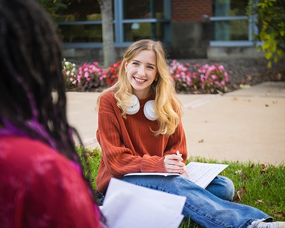 Student sitting on University lawn talking with student in foreground, while smiling. 